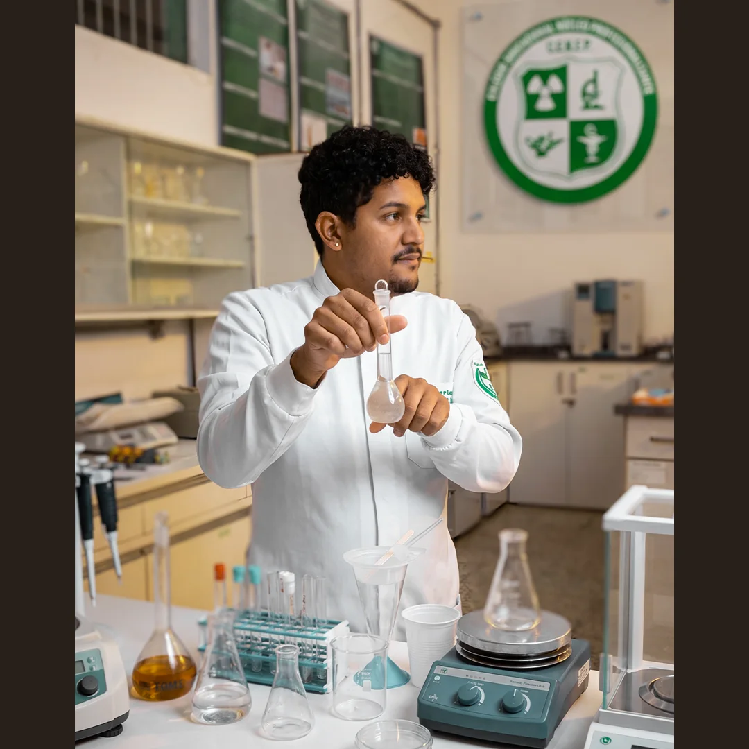 Aluno de jaleco branco manuseia balão volumétrico em laboratório do C.E.N.E.P, com o brasão da escola na parede ao fundo.