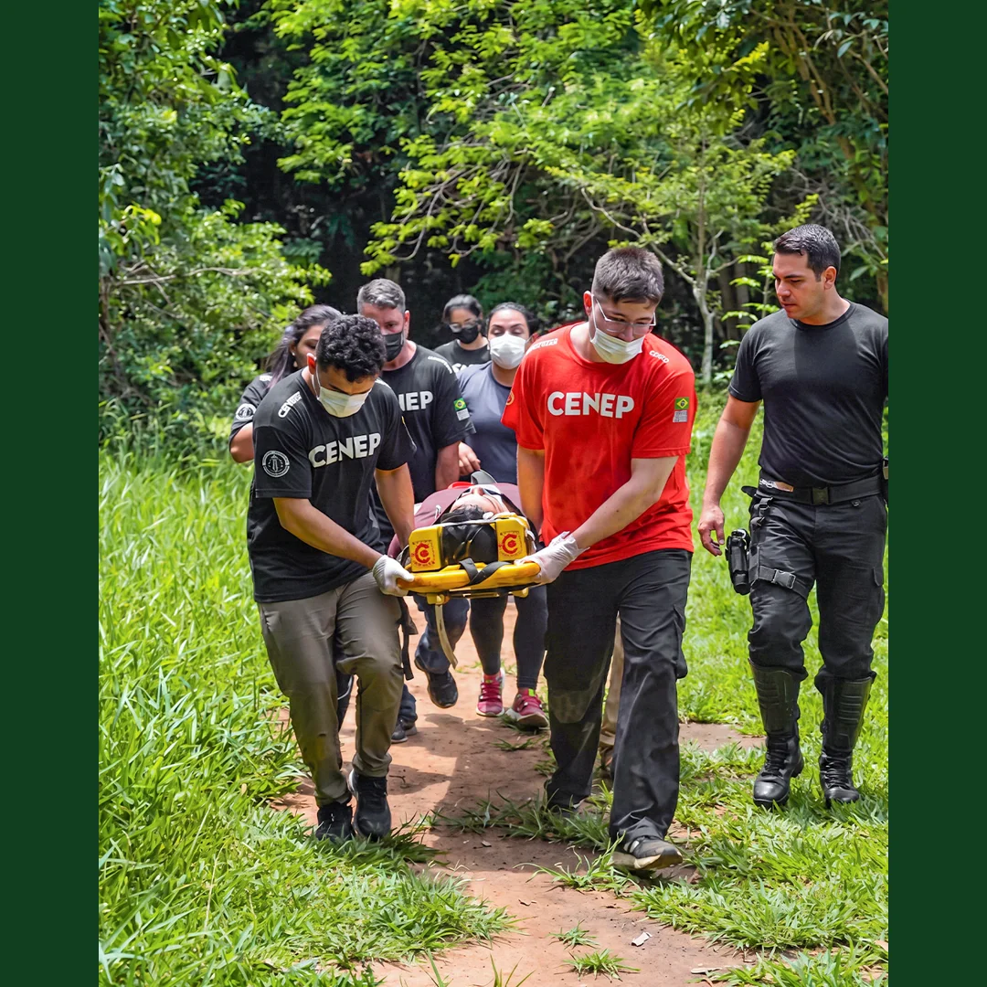 Alunos com camisetas CENEP e máscaras carregam maca rígida amarela com vítima simulada durante treinamento de resgate em trilha.