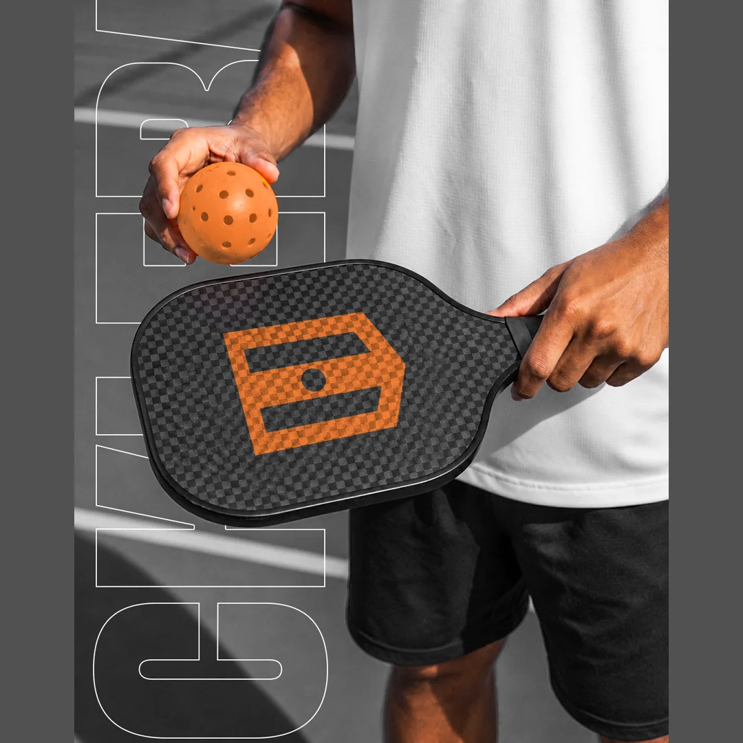 Jogador em quadra segurando raquete de pickleball em fibra de carbono com logo laranja da Hyperlight e bola laranja na outra mão
