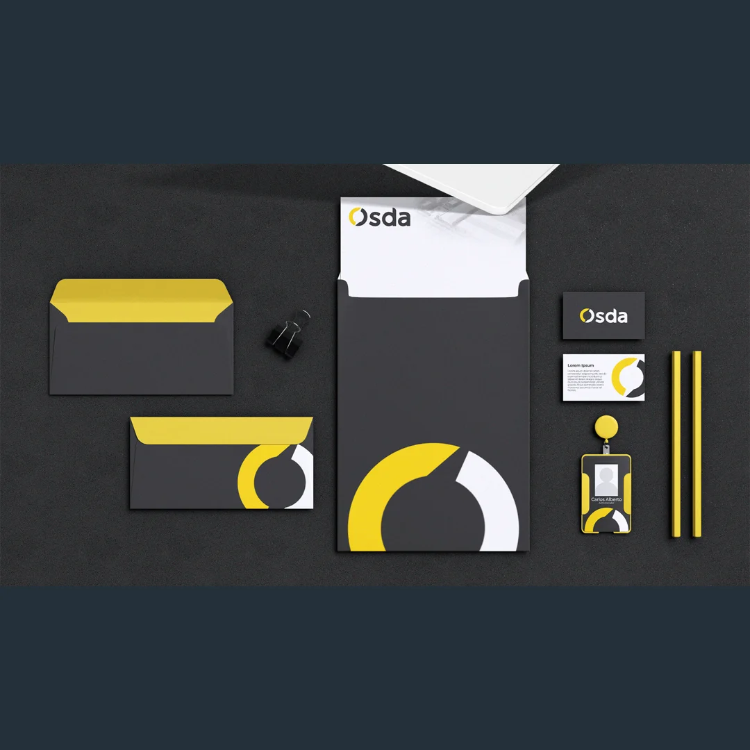 Mockup de papelaria sobre superfície preta texturizada: envelopes grafite com aba amarela, pasta com o símbolo ampliado, papel timbrado, cartões de visita, canetas amarelas e crachá.
