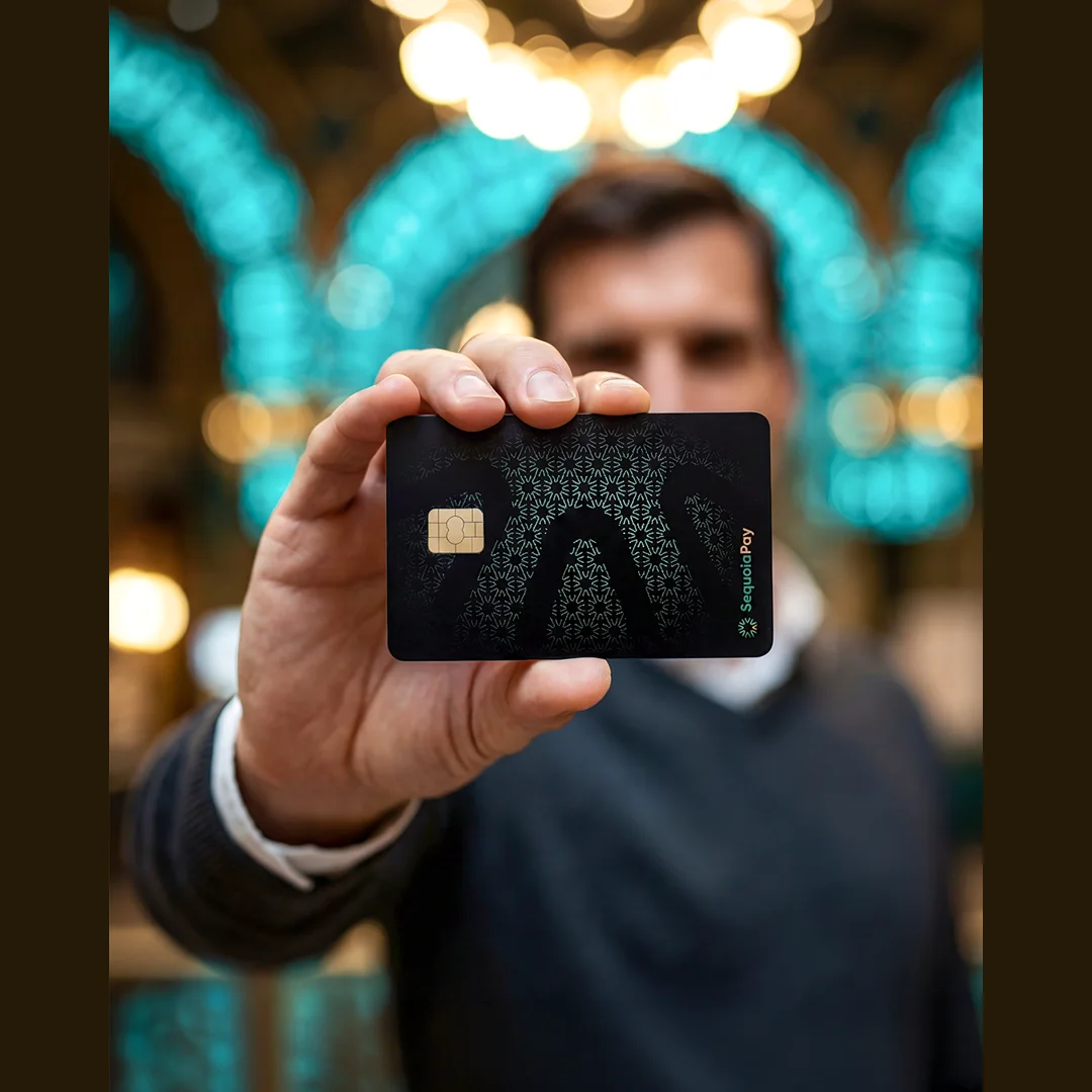 Mão segurando o cartão preto SequoiaPay com padrão gráfico e chip dourado, fundo desfocado com luzes turquesa