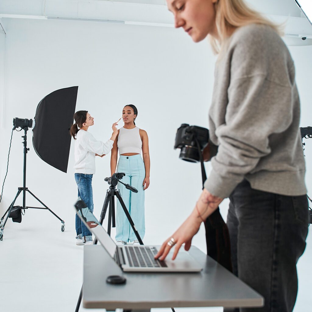 Autoridade visual o que sustenta a percepção de uma marca | Agência Wys | Agência Wys Behind the scenes of a fashion photoshoot in a bright studio makeup artist applying makeup to a model as another photographer adjusts a camera a laptop on a table in the foreground