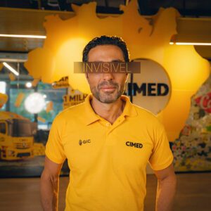 Man in a yellow polo with CIMED logos stands in a yellow themed space A dark band labeled INVISIVEL covers his eyes for the shot